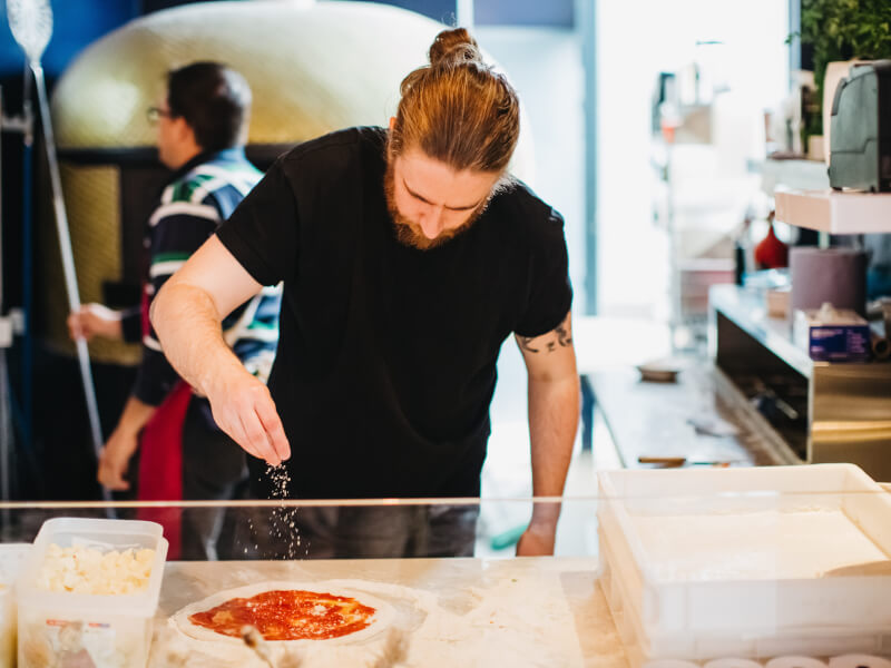 A man sprinkles parmesan onto a dough pizza base with a traditional pizza oven in the background